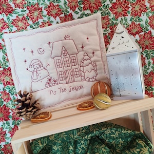 Peut inclure: Un coussin brodé blanc sur le thème de Noël avec une maison, un bonhomme de neige et le texte "Tis The Season". Le coussin est sur une caisse en bois avec un tissu vert à motif de feuilles dorées.