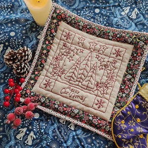 May include: A Christmas-themed embroidered square featuring trees, stars, and snowflakes, with the words "Merry Christmas." The square is bordered with patterned fabric and sits on blue floral fabric. A candle, pine cones, and berries are also present.