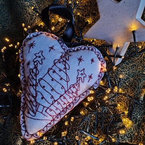 Op de afbeelding: Een wit hartvormig kussen met rode borduursels van een kerstboom en sterren. Het kussen hangt aan een zwart lint en is omgeven door een string met warm witte kerstverlichting.