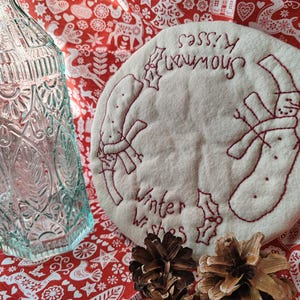 May include: A white fabric circle with red embroidery that says "Sassy & Fabulous" and "Winter Wishes". The fabric circle is on a red background with a white pattern of Scandinavian Christmas designs. There are two pine cones on the red background.