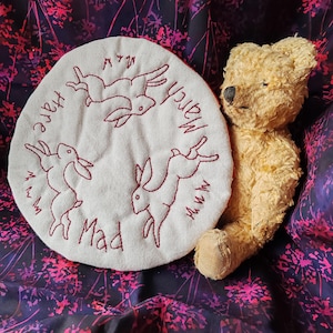Op de afbeelding: Een witte stoffen cirkel met rode borduursels van drie konijnen en de woorden "Mad March Hare". Een bruine teddybeer ligt op de stoffen cirkel.