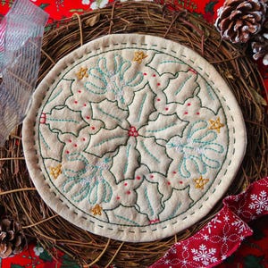 May include: A round, cream-coloured fabric coaster with a green holly leaf design embroidered in a circular pattern. The coaster is surrounded by a brown woven wreath and a red ribbon with white snowflakes.