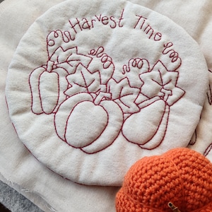 May include: A cream-colored, round quilted trivet with embroidered pumpkins and leaves in a burgundy color. The text "Harvest Time" is embroidered in a circular pattern. An orange crocheted pumpkin is in the foreground, suggesting a fall theme.