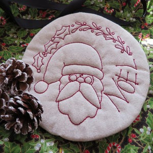 May include: A white fabric ornament with red embroidery of Santa Claus and holly leaves. The ornament says "Noel" in red embroidery.