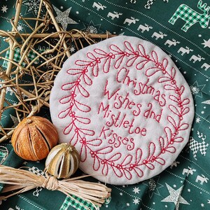 May include: White fabric circle with red embroidery of a wreath and the text "Christmas Wishes and Mistletoe Kisses". The circle is on a green fabric with a white reindeer pattern.