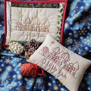 May include: A festive Christmas-themed display featuring a square quilted wall hanging and a rectangular pillow, both embroidered with a town scene and the text "O Little Town." The background is a blue fabric with gold and white snowflakes.