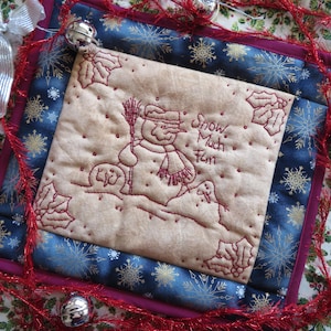 Op de afbeelding: Een gequilte wandhanger met een sneeuwpop en de tekst "Snow Much Fun" in rode borduurwerk. De achtergrond is blauw met gouden sneeuwvlokken en een rode rand.
