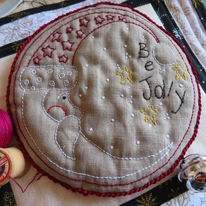 May include: A beige fabric circle with red stitching around the edge. The fabric is embroidered with a white crescent moon, white stars, and the words "Be Jolly" in black thread.