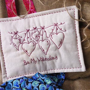 May include: A white fabric wall hanging with red embroidery of a string of hearts and the text "Be My Valentine".