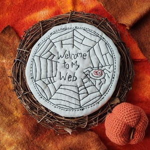 May include: A Halloween-themed decorative piece featuring a white fabric circle embroidered with a spider web design and the text "Welcome to My Web." A small, crocheted orange pumpkin sits nearby, all set within a woven wreath.