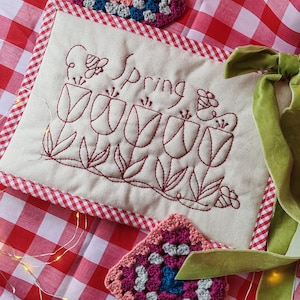 May include: A cream-coloured embroidered cushion featuring the word "Spring" and tulip designs in red thread. The cushion is bordered with red and white gingham fabric. Two crocheted squares and a green velvet ribbon are also in the image.