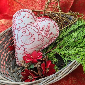 May include: A white heart-shaped cushion with red embroidery of a bird singing in a snowy forest. The cushion is in a wicker basket with red poinsettia flowers and green pine branches.