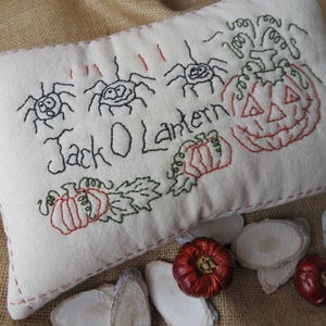 May include: A white pillow with red stitching and embroidered details. The pillow features a jack-o-lantern, pumpkins, and spiders with the text "Jack O Lantern".