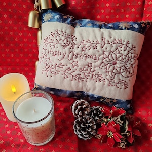 Könnte beinhalten: Ein cremefarbenes Weihnachtskissen mit roter Stickerei "Happy Christmas" und blauem Rand. Das Bild zeigt festliche Dekorationen: Kerzen, Tannenzapfen und Messingglocken, alles vor einem roten Hintergrund mit Muster.