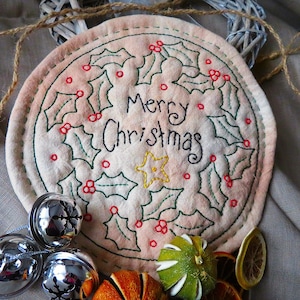 May include: A white fabric circle with green holly leaves and red berries embroidered around the words "Merry Christmas".