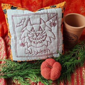 Pode incluir: Uma almofada branca com um rosto de gato bordado a vermelho e a palavra "Catoween" nela. A almofada está sobre um tecido vermelho e laranja com uma pequena abóbora de crochê laranja à frente.