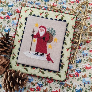 May include: A cross-stitch Christmas scene depicting Santa Claus in a red coat, holding a staff and a sack of gifts, with a red bird. The artwork is set on a light beige fabric with a scalloped edge, surrounded by a green and red border, and placed on a festive fabric with a winter village pattern.