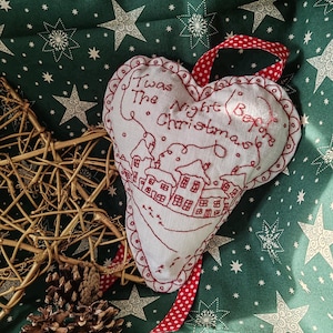 Pode incluir: Adorno de tecido branco em forma de coração com fita de bolinhas vermelhas. O adorno é bordado com o texto "Twas The Night Before Christmas" e uma pequena cena de vila.