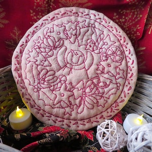 May include: A white fabric Christmas ornament with red embroidery depicting a wreath of holly, berries, and stars. The ornament is sitting in a wicker basket with two white tea lights.