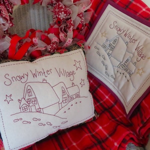 May include: Two embroidered pillow covers with the text "Snowy Winter Village" and a design of houses and snow. The pillow covers are on a red and white plaid blanket.
