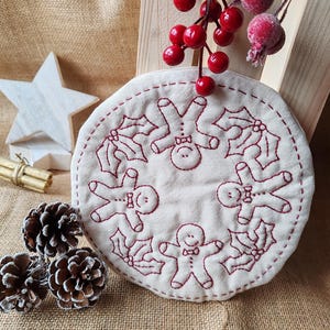 May include: A round, white fabric ornament with red embroidery of gingerbread men and holly leaves. The ornament is surrounded by pine cones and a wooden crate with red berries.