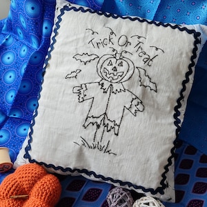 May include: A white pillow with a black embroidered pumpkin and the text "Trick Or Treat". The pillow is on a blue and purple patterned fabric with a crocheted orange pumpkin and white decorative balls.