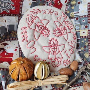 May include: A white fabric ornament with three embroidered angels and the text "Peace on Earth" in red thread. The ornament is surrounded by dried orange slices, nuts, and a nutcracker on a red and gray patterned fabric.