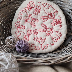 May include: A white fabric circle with red embroidery of bunnies, eggs, and the text "Hoppy Easter". The circle is in a wicker basket with white and purple decorative balls.