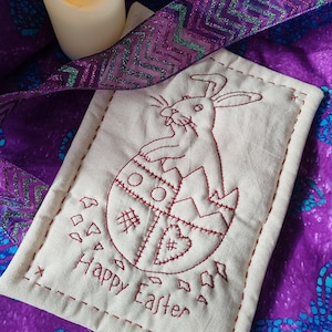 May include: A cream-coloured, rectangular Easter-themed fabric item with a stitched design of a bunny emerging from a cracked egg. The words "Happy Easter" are stitched below. A purple ribbon and a candle are in the background.