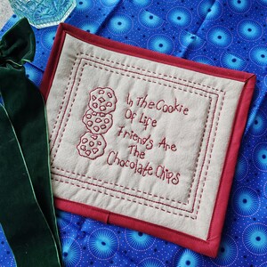 May include: A beige quilted coaster with a red border and embroidered text that reads "In the Cookie Of Life Friends Are The Chocolate Chips".