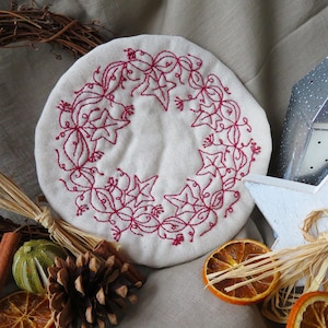 Può includere: Un cerchio di tessuto bianco con un disegno ricamato rosso a forma di ghirlanda di foglie e stelle. Il tessuto è circondato da elementi naturali come bastoncini di cannella, frutta secca e pigne.