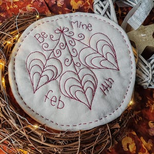 May include: A circular, off-white fabric coaster featuring red embroidered hearts and the words "Be Mine feb 14th". The coaster is set within a brown twig wreath with fairy lights.