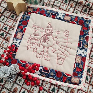 May include: A square fabric panel with a beige center featuring red embroidered text that reads snowman For Sale assembly Required, a snowman, and a tree. The panel is bordered by a blue fabric with red and white holiday patterns.