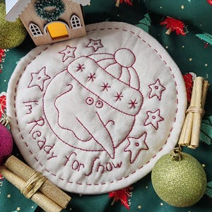 May include: A circular, cream-coloured fabric coaster featuring a burgundy embroidered snowman design and the text "I'm Ready for Snow". The coaster is surrounded by Christmas decorations, including baubles and cinnamon sticks.