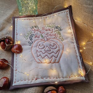 May include: A square, cream-coloured fabric item with a brown border features an embroidered jack-o'-lantern and the words "Give Thanks for All Things" in orange and green thread. Decorative lights and conkers surround the item.