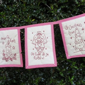 May include: A red and white gingham banner with five white fabric flags. Each flag has a different Christmas-themed design and text. The text on the flags reads "Peace On Earth", "Oh Come All Ye Faithful", "Oh Christmas Tree", and two other phrases that are not fully visible in the image.