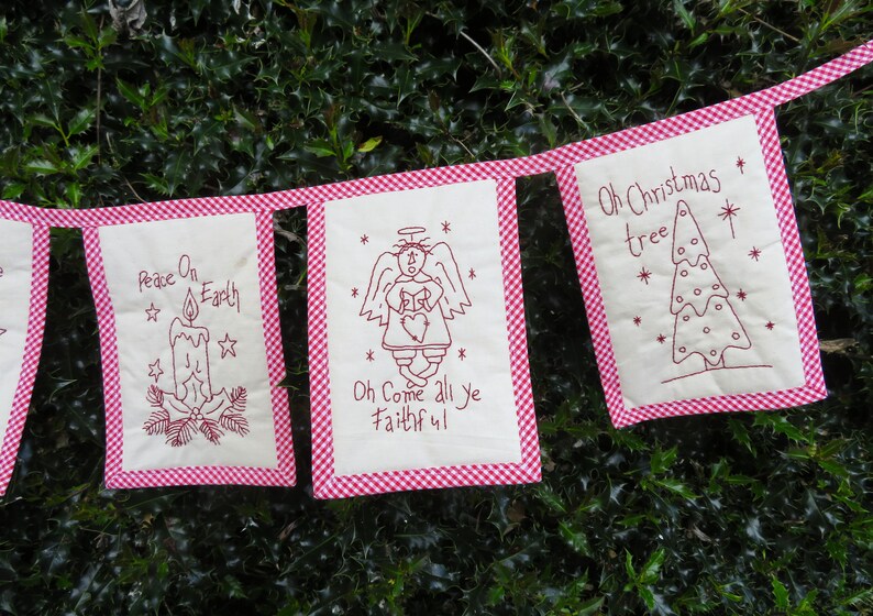 May include: A red and white gingham banner with five white fabric flags. Each flag has a different Christmas-themed design and text. The text on the flags reads "Peace On Earth", "Oh Come All Ye Faithful", "Oh Christmas Tree", and two other phrases that are not fully visible in the image.