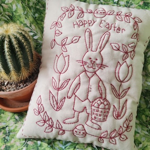 May include: Cream-colored Easter pillow with red embroidered design. Features a bunny holding a basket of eggs, surrounded by a floral wreath and tulips. The words "Happy Easter" are embroidered above the bunny.