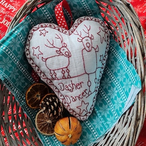 May include: A white heart-shaped ornament with red embroidery of two reindeer and the text "Dasher Dancer". The ornament is in a wicker basket with a teal and white patterned fabric.