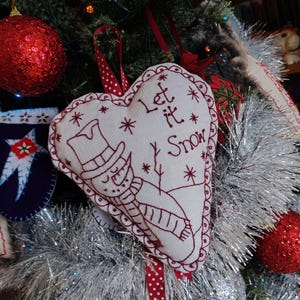 May include: A white heart-shaped felt ornament with red stitching and the text "Let it Snow" and a snowman design. The ornament is hanging on a Christmas tree with red and white polka dot ribbon.