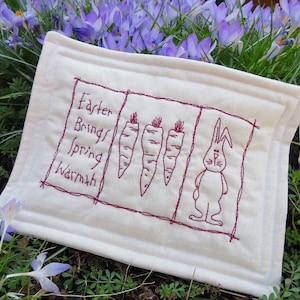 May include: White fabric with red embroidery of a bunny and carrots. The text reads "Easter Brings Spring Warmth".