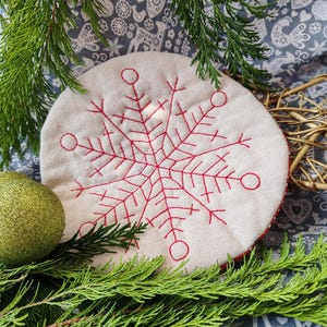 May include: A round, white fabric ornament with a red embroidered snowflake design. The ornament is surrounded by green pine branches.