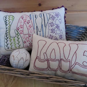 May include: Three embroidered cushion covers with the word "LOVE" spelled out in different styles. The cushions are in a wicker basket.