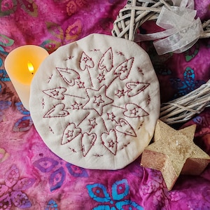 May include: A cream-coloured, round embroidered item with a star and heart design in red thread. A lit candle, a gold star, and a woven wreath with a white ribbon sit on a pink and purple floral patterned fabric.