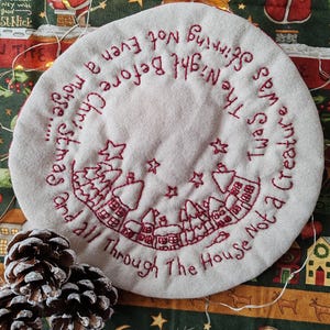 May include: A round, off-white Christmas tree skirt with red embroidered text and a winter village scene. The text reads "Twas the Night Before Christmas..." and "And all Through The House Not a Creature". Two pine cones are in the foreground.