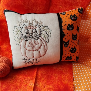 May include: A Halloween cushion featuring an embroidered cat in a pumpkin design. The cushion has a cream front with black trim and an orange side patterned with black cat faces. The word "FALL" is embroidered below the cat.