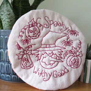 May include: A white fabric circle with red embroidery depicting a witch's hat, pumpkins, and spiders. The text "Spooky Witchy Good" is embroidered on the fabric.