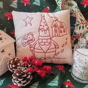 Op de afbeelding: Een vierkant, beige kussen geborduurd met een rood kerstmanontwerp, een ster, een huis en de woorden "Merry Christmas". Het kussen ligt op groene stof met kerstboompatronen, samen met dennenappels, rode bessen en een kaars.