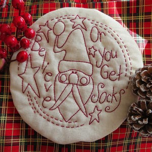 May include: A white fabric circle with red embroidery depicting a gnome with a hat and the text "Believe or you get socks". The circle is surrounded by a red and black tartan fabric.