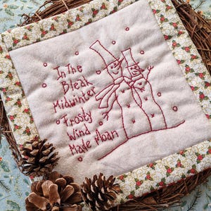May include: A white fabric square with a red embroidered design of two snowmen and the text "In the Bleak Midwinter Frosty Wind Made Moan". The square is framed with a gold and red patterned fabric border.
